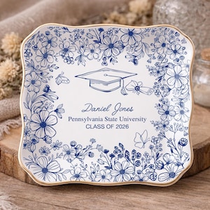 May include: A white ceramic plate with a gold rim, adorned with blue floral patterns and a graduation cap illustration. The plate is personalized with the name "Daniel Jones", "Pennsylvania State University", and "CLASS OF 2026".
