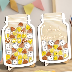 May include: Two wooden reward jars shaped like mason jars, each with a white outline and wooden stand. The jars are filled with cartoon images of poop, toilet paper, and yellow shapes. The text "Annie's Reward Jar" is written on the jars.