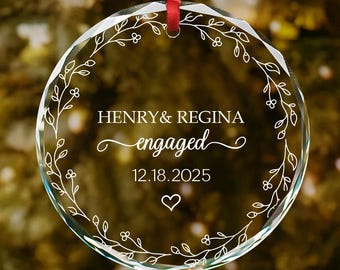 May include: A clear, round glass ornament with a red ribbon for hanging. The ornament features the names "HENRY & REGINA", the word "engaged", the date "12.18.2025", and a heart symbol. A floral design encircles the text.