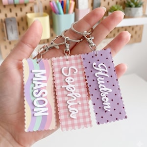 Puede incluir: Tres llaveros rectangulares de acrílico con bordes festoneados y cierres de mosquetón de metal plateado. El primero presenta un patrón de arcoíris pastel con el nombre MASON, el segundo un patrón de cuadros vichy rosa con Sophia y el tercero estrellas moradas con Hudson.