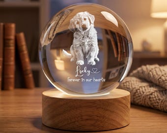 May include: A clear glass sphere featuring an etched image of a puppy sitting down. Below the puppy, the text reads "Lucky" with a heart symbol and "Forever in our hearts". The sphere is mounted on a round, light-colored wooden base.