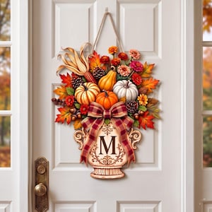 May include: A decorative door hanging featuring a cream-colored urn with the letter M, filled with orange, white, and yellow pumpkins, pinecones, corn, and autumn leaves. A plaid ribbon is tied around the urn, and a rope hanger is attached.