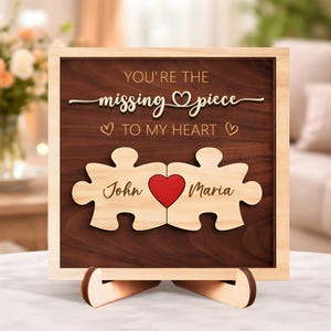May include: Wooden puzzle piece sign with the text "You're the missing piece to my heart." The sign features two puzzle pieces with the names "John" and "Maria" and a red heart. The sign is displayed on a wooden stand.