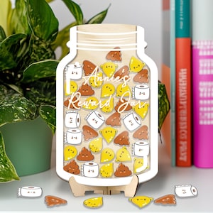 Könnte beinhalten: Ein hölzernes Belohnungsglas mit dem Text "Annie's Reward Jar" in Schreibschrift. Das Glas ist mit Holzmarken in Form von Kack-Emojis, Zitronenscheiben und Toilettenpapierrollen gefüllt. Das Glas steht auf einem Holzständer.