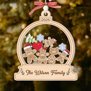 May include: A wooden Christmas ornament shaped like a snow globe, featuring gingerbread people with names, snowflakes, and a heart. The ornament has the year 2025 and the text "The Wilson Family". It is decorated with a red and white ribbon.