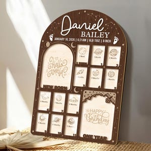 May include: A wooden milestone board with the name "Daniel Bailey" and birth details. It features spaces for photos and milestones from newborn to one year, with the text "Hello World" and "Happy Birthday".