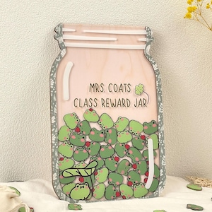 Puede incluir: Un tablero de recompensas de clase de madera en forma de tarro con el texto "MRS. COATS CLASS REWARD JAR". El tarro está lleno de fichas de rana verdes, cada una con una baya roja. Fichas de rana adicionales están esparcidas.
