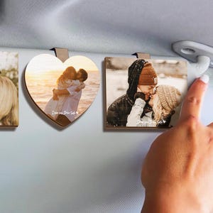 May include: Two wooden photo car air freshener ornaments, one heart-shaped and one rectangular. The heart-shaped ornament features a photo of a couple embracing on a beach at sunset. The rectangular ornament features a photo of a couple kissing in the snow. Both ornaments have the text "I love you, Drive Safe" printed on them.