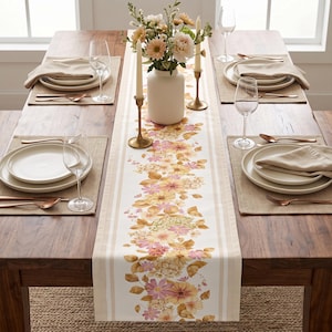 Puede incluir: Un camino de mesa blanco con un patrón floral central de flores y hojas rosas, amarillas y naranjas, flanqueado por rayas beige. El camino está colocado sobre una mesa de comedor de madera con cuatro servicios que incluyen platos, servilletas, vasos y cubiertos de color cobre.