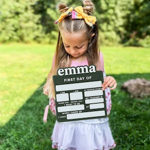 May include: A dark green rectangular sign with white text reading "emma" and "FIRST DAY OF" followed by blank fields for "I AM" years old, "I AM" inches tall, "MY FAVORITE COLOR IS", "MY TEACHER IS", "I WANT TO BE" when I grow up, "AT", and "I LOVE".