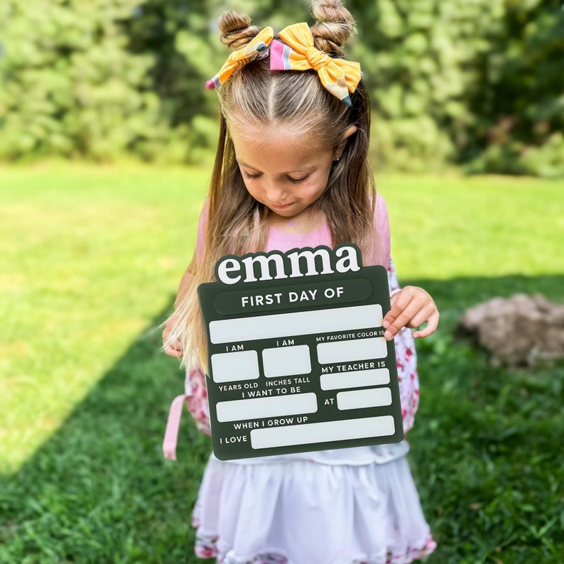 May include: A dark green rectangular sign with white text reading "emma" and "FIRST DAY OF" followed by blank fields for "I AM" years old, "I AM" inches tall, "MY FAVORITE COLOR IS", "MY TEACHER IS", "I WANT TO BE" when I grow up, "AT", and "I LOVE".