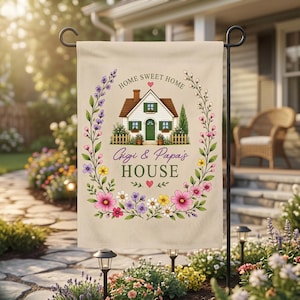 Könnte beinhalten: Eine beige Gartenflagge mit einer zentralen Abbildung eines weißen Hauses mit grüner Tür und braunem Dach. Der Text lautet HOME SWEET HOME und Gigi & Papa's HOUSE. Ein Blumenkranz aus rosa, gelben und violetten Blüten umrahmt das Haus.