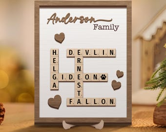 May include: A wooden frame with a white background and a Scrabble tile crossword puzzle that spells out "Anderson Family". The puzzle includes a paw print tile and hearts.