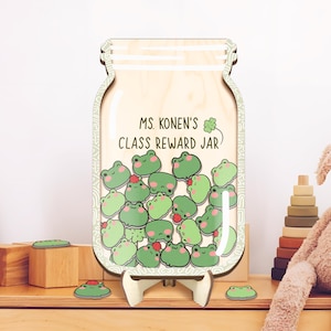 Puede incluir: Un tarro de recompensa de clase de madera con forma de frasco de vidrio con un trébol verde y el texto "Ms. Konen's Class Reward Jar". El tarro está lleno de fichas con forma de rana verde.