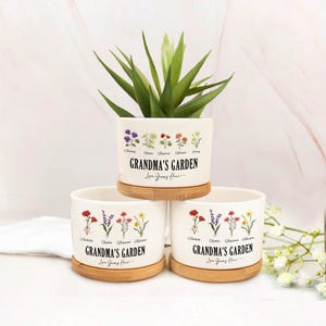 May include: Three white ceramic flower pots with bamboo saucers. Each pot is decorated with floral illustrations and the text "GRANDMA'S GARDEN Love Grows Here." One pot holds a green succulent plant.