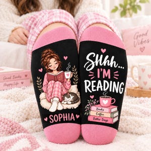 May include: A pair of black socks with pink cuffs. The left sock features an illustration of a person reading a book with a cat and the name Sophia. The right sock displays the text Shhh... I'M READING above a stack of books and a coffee mug.