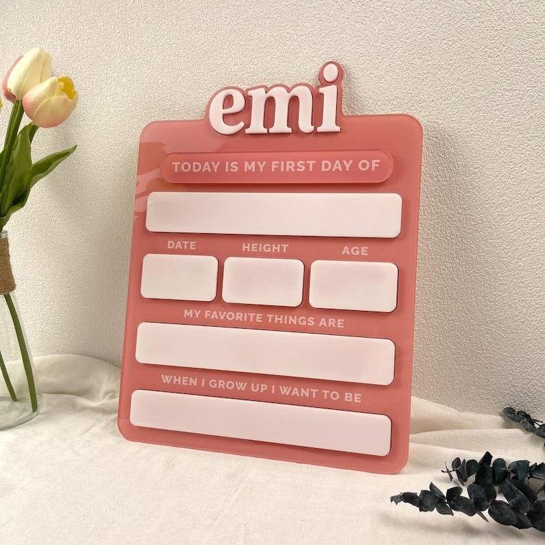 May include: A pink acrylic milestone board featuring the name emi in raised white lettering. The board includes printed text fields for Today Is My First Day Of, Date, Height, Age, My Favorite Things Are, and When I Grow Up I Want To Be.