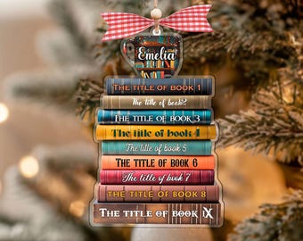 Puede incluir: Adorno navideño personalizado con una pila de libros, cada uno con un título. El adorno tiene un lazo a cuadros rojo y blanco y el nombre "Emelia". El adorno está colgado de un árbol de Navidad.