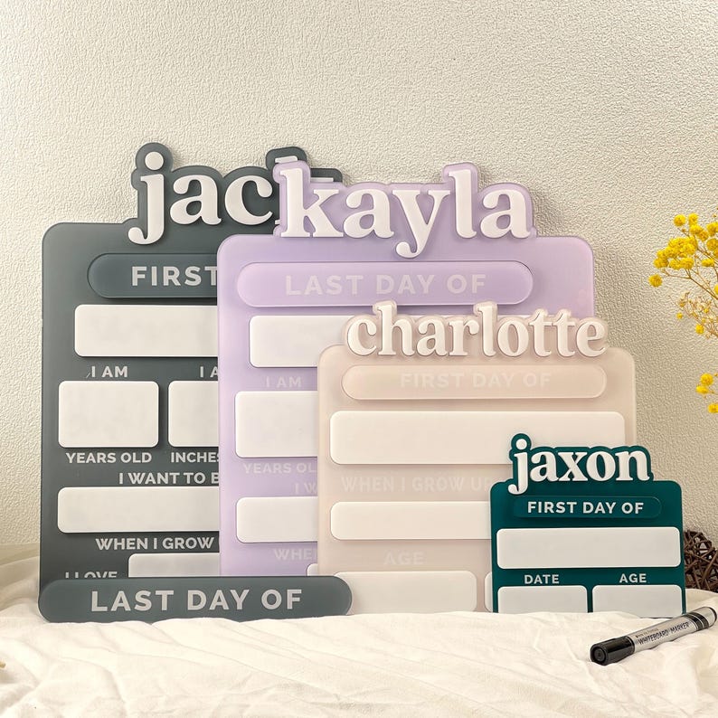 May include: Four rectangular acrylic milestone boards in dark gray, lavender, beige, and dark green. Each board features a raised name at the top, including "jac", "kayla", "charlotte", and "jaxon", with blank white spaces for writing details like dates, ages, and goals.