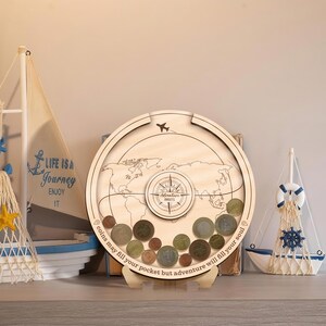 May include: A circular wooden coin bank with a clear front panel displaying a world map, a compass graphic, and an airplane icon. The text reads "Adventure AWAITS" and "coins may fill your pocket but adventure will fill your soul."