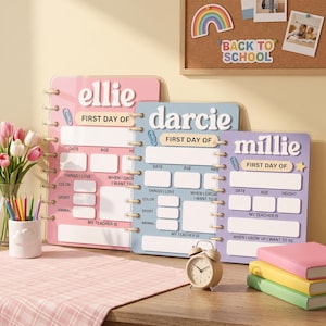 Pode incluir: Três quadros brancos de acrílico para marcos escolares, personalizados com os nomes Ellie, Darcie e Millie. Cada quadro apresenta secções para data, idade, altura, professor e interesses, com um design em espiral e cores pastel.