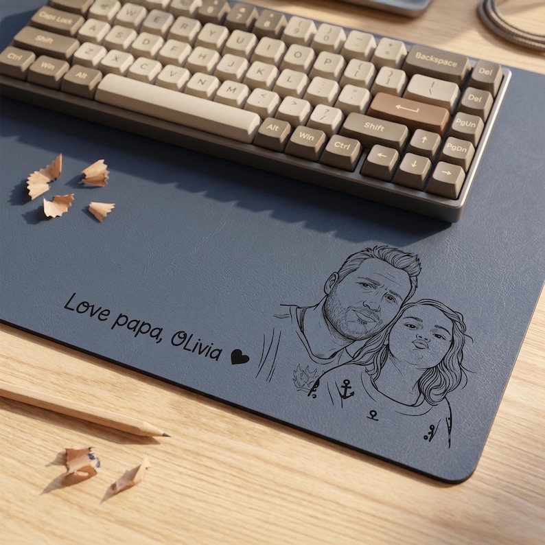 Peut inclure: Un clavier m&eacute;canique gris et beige est pos&eacute; sur un tapis de bureau bleu. Le tapis pr&eacute;sente une illustration en noir et blanc d'un p&egrave;re et d'un enfant, avec le texte "Love papa, Olivia" et un symbole de c&oelig;ur. Des copeaux de crayon sont &eacute;parpill&eacute;s.