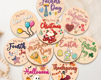 May include: A collection of round wooden plaques with holiday-themed designs. Each plaque features the phrase "My First" followed by a holiday name, such as "Christmas" or "Valentine's Day." The plaques are decorated with festive illustrations.