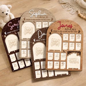 May include: Four wooden baby milestone boards with arched tops. Each board has a photo frame, the words "Hello World" and "New Born". One board says "Happy Birthday" and "One Year". The boards are in shades of brown.