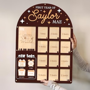 May include: A dark brown wooden baby milestone board with the text "First Year of Saylor Mae". It has spaces for photos and details like date, time, weight, and length. The board includes the text "Welcome to the World" and "New Born".