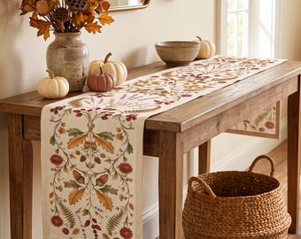 May include: A long, rectangular table runner featuring a cream-colored fabric base with a printed botanical pattern. The design includes illustrations of red flowers, brown acorns, green oak leaves, ferns, and purple sprigs arranged in a symmetrical motif.