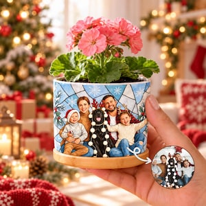 May include: A cylindrical ceramic planter with a bamboo base, featuring a printed illustration of a family and a black dog wearing reindeer antlers. A pink geranium plant grows from the top. A small circular inset shows the original photo.