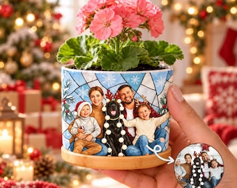 May include: A cylindrical ceramic planter with a bamboo base, featuring a printed illustration of a family and a black dog wearing reindeer antlers. A pink geranium plant grows from the top. A small circular inset shows the original photo.