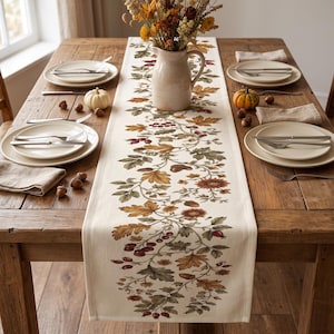 May include: A cream-colored table runner featuring a printed botanical pattern of autumn leaves, acorns, and red berries. The runner is centered on a wooden dining table set with white plates, silver cutlery, linen napkins, and a ceramic pitcher.