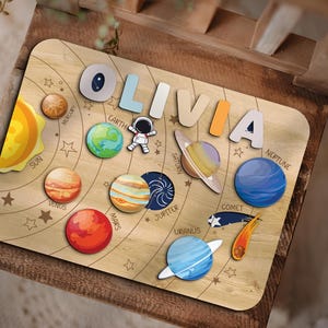 Könnte beinhalten: Ein Holzpuzzle mit den Planeten unseres Sonnensystems. Die Puzzleteile sind in Form der Planeten gestaltet und tragen die Namen der einzelnen Planeten. Das Puzzle enthält auch einen Kometen und einen Astronauten. Das Puzzle ist mit dem Namen "Olivia" personalisiert.