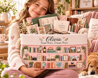May include: A rectangular pink fabric storage bin featuring a printed illustration of three bookshelves filled with books, plants, and decorative items. The bin is personalized with the name Olivia and the text Read More, Dream More.