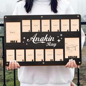 Puede incluir: Un tablero de graduación de madera personalizado con espacios para fotos desde preescolar hasta 11º grado. El tablero es de color marrón oscuro con detalles en madera clara y presenta el nombre "Anakin Ray" en una fuente decorativa, junto con una ilustración de birrete.