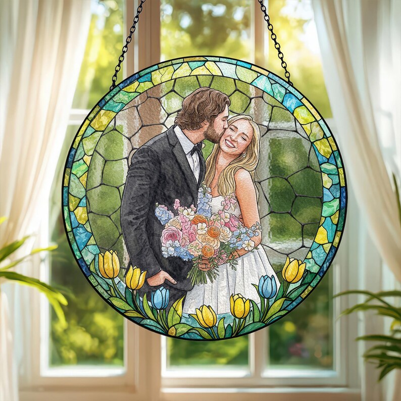 May include: A round stained glass window featuring a couple on their wedding day. The man in a black suit kisses the woman in a white dress. The window is framed with colorful flowers and a chain for hanging.