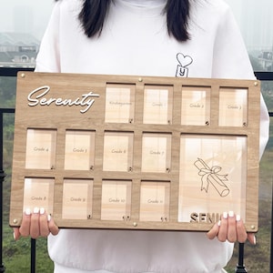 Puede incluir: Un marco de fotos de madera con 12 ranuras para fotos, etiquetadas con los grados de Kindergarten a Grado 11. El marco está grabado con la palabra "Serenity" y la palabra "Senior" en la parte inferior.