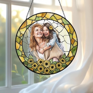 May include: Round stained glass window art featuring a mother and child embracing, surrounded by sunflowers. The artwork is framed with a black metal chain and a yellow and green stained glass border. The image depicts a joyful moment.