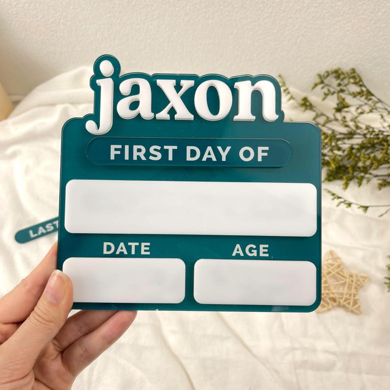 May include: A dark teal acrylic sign featuring the white text Jaxon and FIRST DAY OF. The sign includes a large blank white rectangular space for writing, followed by two smaller blank white rectangular spaces labeled DATE and AGE.