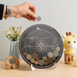 May include: A circular wooden coin bank with a clear front panel featuring a world map, a compass graphic, and the text "Adventure Awaits" and "coins may fill your pocket but adventure will fill your soul" with a small airplane icon.