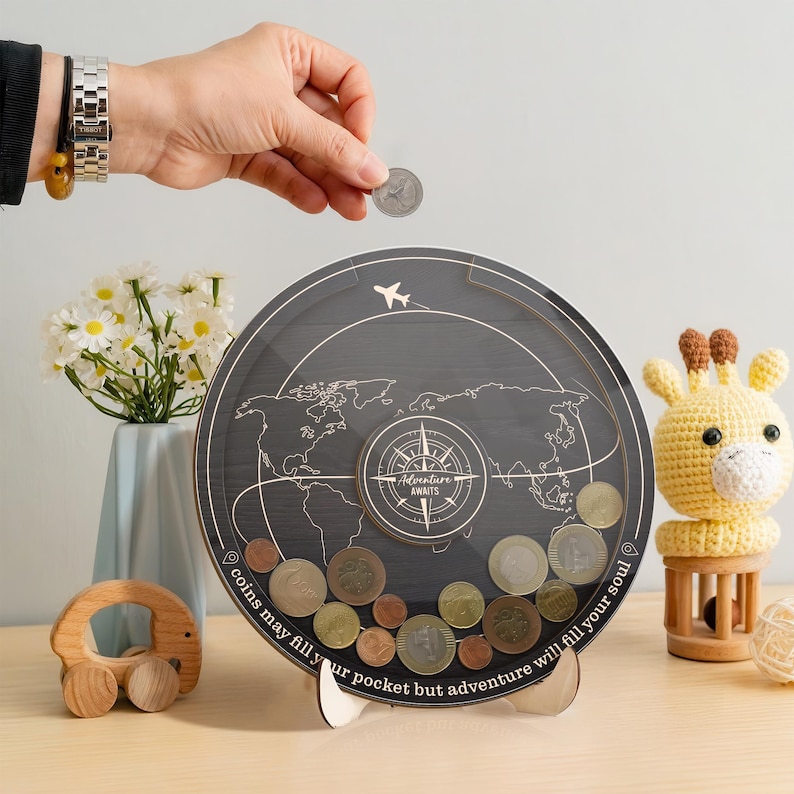 May include: A circular wooden coin bank with a clear front panel featuring a world map, a compass graphic, and the text "Adventure Awaits" and "coins may fill your pocket but adventure will fill your soul" with a small airplane icon.