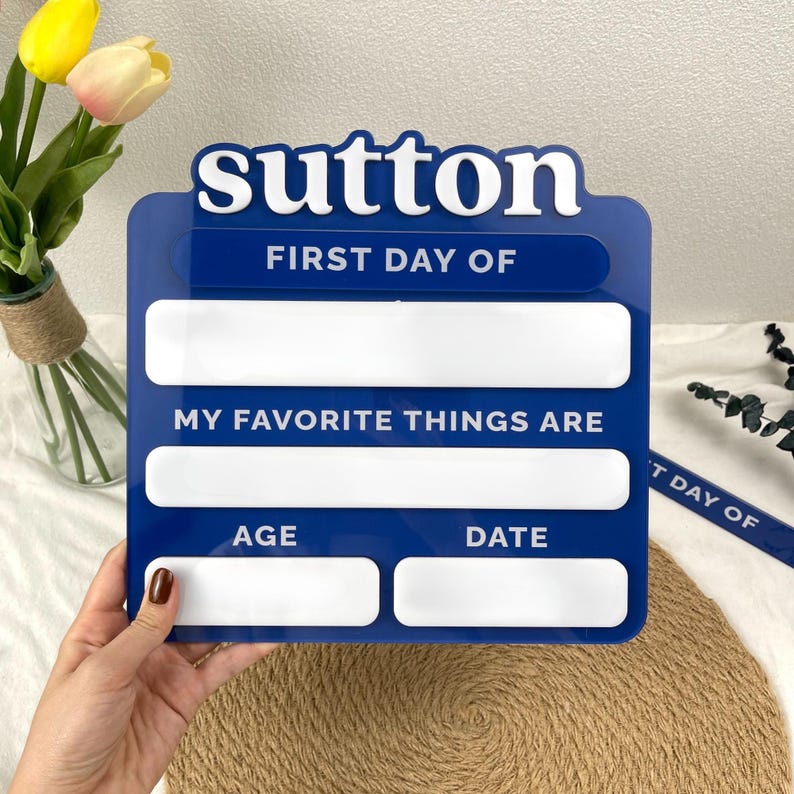 May include: A blue and white acrylic sign featuring the name sutton in large letters at the top. The sign includes the text FIRST DAY OF, MY FAVORITE THINGS ARE, AGE, and DATE, with blank white rectangular spaces for writing information.
