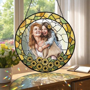 May include: Round stained glass window art featuring a mother and child embracing, surrounded by a yellow and green border. The bottom edge is decorated with yellow sunflowers. The artwork is suspended by a chain.