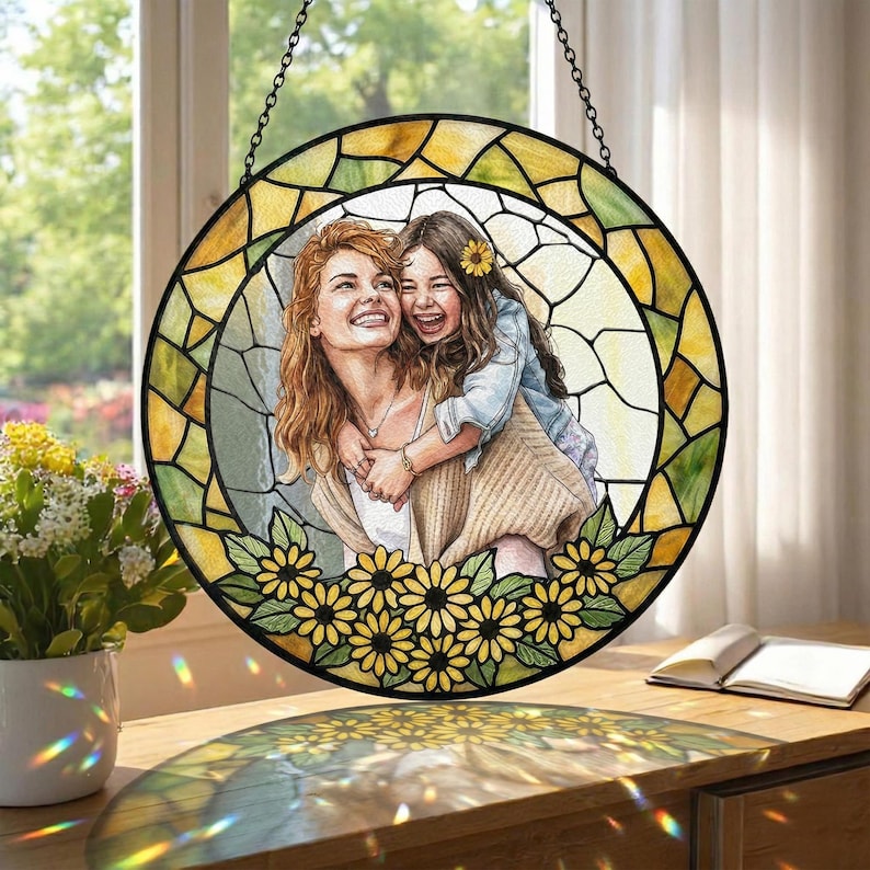 May include: Round stained glass window art featuring a mother and child embracing, surrounded by a yellow and green border. The bottom edge is decorated with yellow sunflowers. The artwork is suspended by a chain.