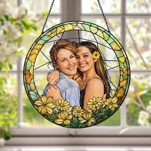May include: Round stained glass window art featuring two women embracing, surrounded by yellow flowers. The artwork has a black chain for hanging. The glass is segmented with yellow, green, and clear panels, creating a vibrant, decorative piece.