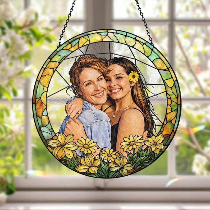 May include: Round stained glass window art featuring two women embracing, surrounded by yellow flowers. The artwork has a black chain for hanging. The glass is segmented with yellow, green, and clear panels, creating a vibrant, decorative piece.