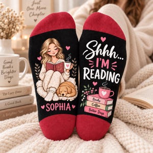 May include: A pair of black socks with red heels and toes. One sock features an illustration of a person reading a book with a dog and the name Sophia. The other sock displays the text Shhh... I'M READING above a stack of books and a coffee mug.