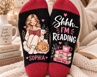 May include: A pair of black socks with red heels and toes. One sock features an illustration of a person reading a book with a dog and the name Sophia. The other sock displays the text Shhh... I'M READING above a stack of books and a coffee mug.