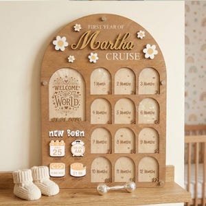 Può includere: Lavagna in legno per i traguardi del bambino con il nome "Martha Cruise" e decorazioni a margherita. Include spazi per foto mensili, una targa "Benvenuti al mondo" e dettagli come data, ora, peso e altezza.